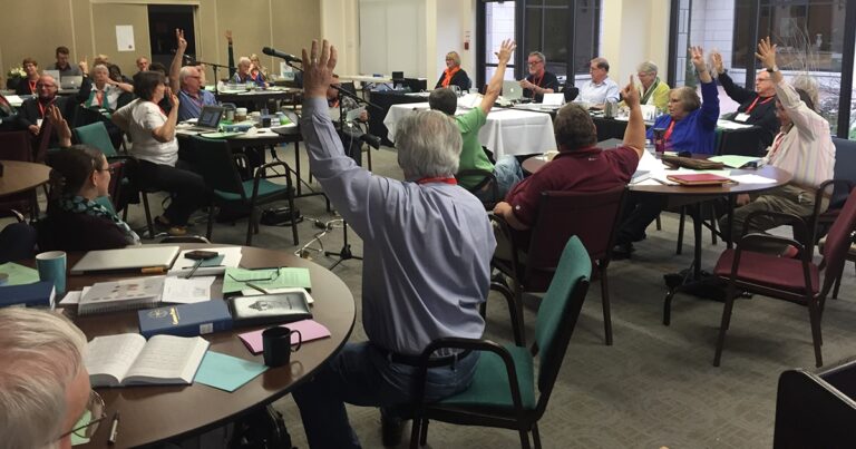 A group of people in a meeting room raising their hands to vote or express agreement, with tables set up for discussion and various materials scattered on the tables.