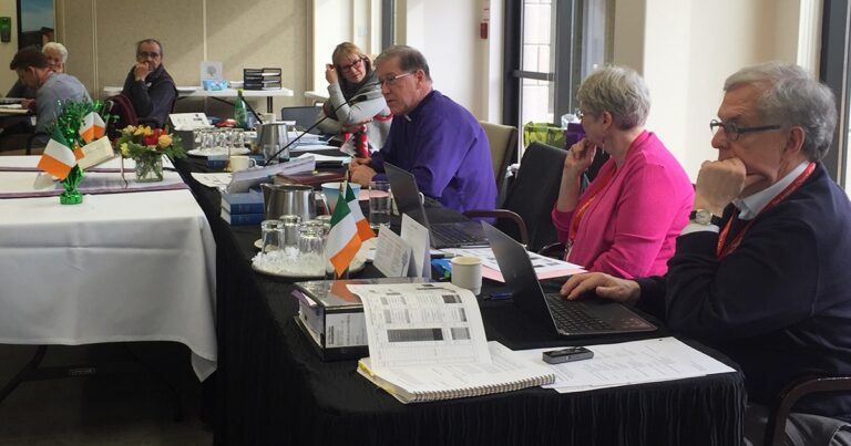Group of professionals engaged in a meeting at a conference table, with laptops, documents, and refreshments visible. An Irish flag is displayed among the materials, indicating a possible connection to Ireland.