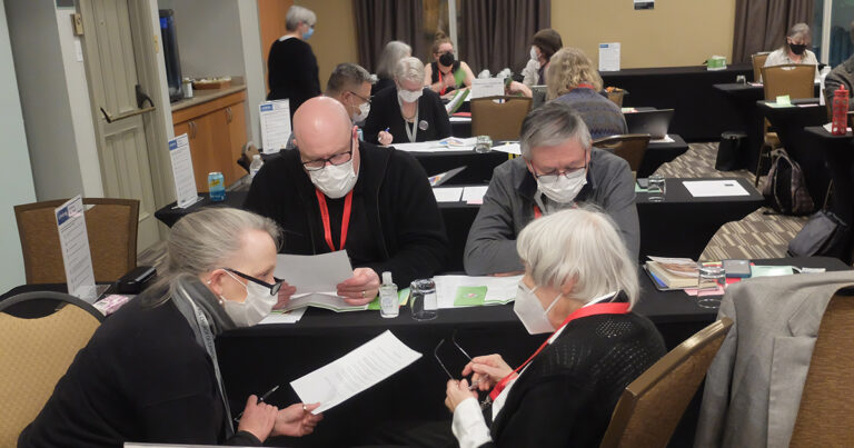 Group of four individuals engaged in discussion at a conference table, reviewing papers and using mobile devices, while wearing masks and lanyards.