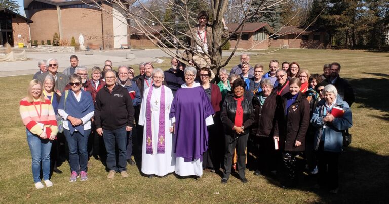 Group photo of a diverse gathering of individuals, including clergy in white and purple vestments, standing outdoors in front of a tree, with buildings visible in the background.