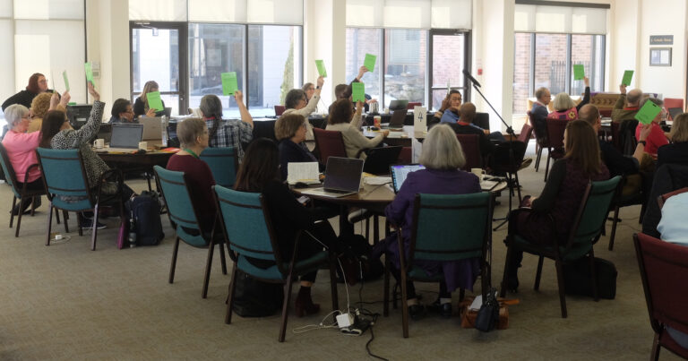 Members of the Council of General Synod seated at tables hold up green voting cards during a session, participating in a formal decision process.