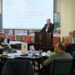 A speaker addresses the Council of General Synod from a podium while members seated at round tables listen and review documents. A presentation slide behind him reads a motion concerning editorial board recommendations and journalistic policies.