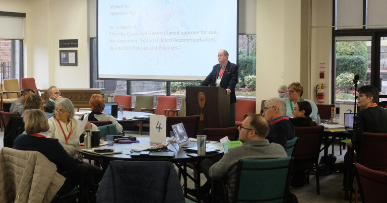 A speaker addresses the Council of General Synod from a podium while members seated at round tables listen and review documents. A presentation slide behind him reads a motion concerning editorial board recommendations and journalistic policies.