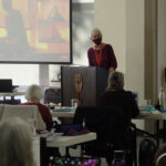 A speaker presenting at a podium in a conference setting, with an audience of older adults seated at tables, engaged in the presentation. A large screen displays visuals related to the talk.