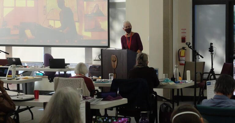 A speaker presenting at a podium in a conference setting, with an audience of older adults seated at tables, engaged in the presentation. A large screen displays visuals related to the talk.