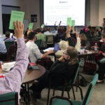 Audience members raising green cards during an interactive session at a conference, with a presentation screen visible in the background.
