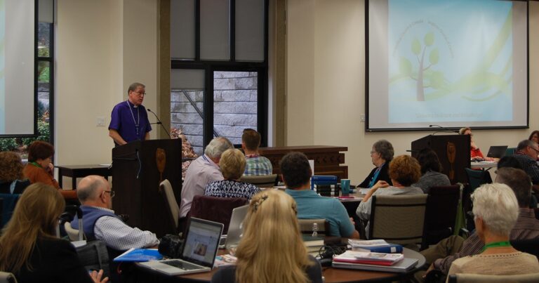A speaker presenting at a podium during a conference, with an audience engaged in the background. The presentation includes a visual aid displayed on a screen.