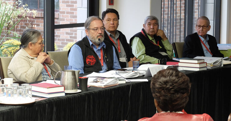 Panel discussion featuring three men seated at a table, engaged in conversation, with one man wearing glasses and a vest, while another man in the background appears attentive.