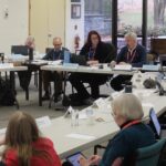 A large committee-style meeting is underway in a bright room with floor-to-ceiling windows. About twenty people sit around several joined tables, laptops open, documents spread out, and nameplates in front of them. Most wear red or blue lanyards. Some participants are speaking while others listen, take notes, or look at screens. Coats and bags rest at their feet. The scene has the focused, slightly weary energy of a full-day governance session, with late-autumn trees visible outside through the windows.