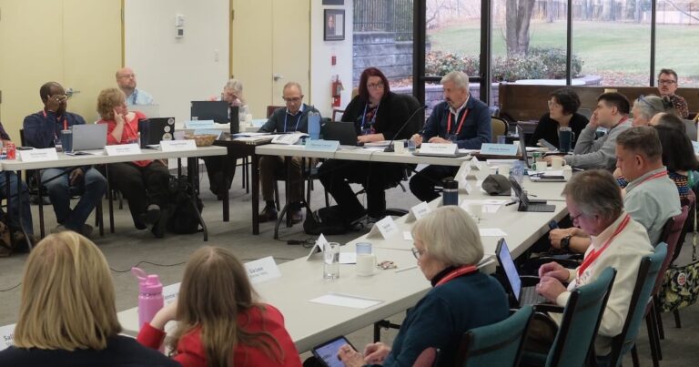 A large committee-style meeting is underway in a bright room with floor-to-ceiling windows. About twenty people sit around several joined tables, laptops open, documents spread out, and nameplates in front of them. Most wear red or blue lanyards. Some participants are speaking while others listen, take notes, or look at screens. Coats and bags rest at their feet. The scene has the focused, slightly weary energy of a full-day governance session, with late-autumn trees visible outside through the windows.