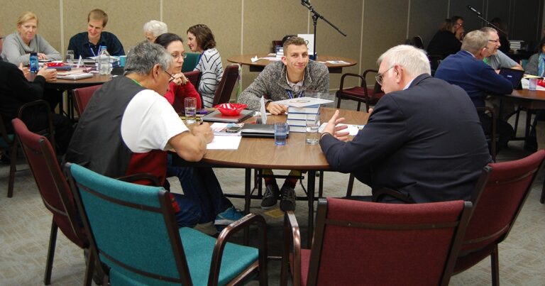 Group discussion at a round table featuring four individuals engaged in conversation, with books and glasses of water present. The setting appears to be a meeting or workshop environment.