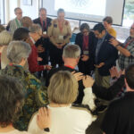 A diverse group of people gathered in a circle, with some individuals bowing their heads and others placing hands on shoulders, engaged in a moment of reflection or prayer during a community event.