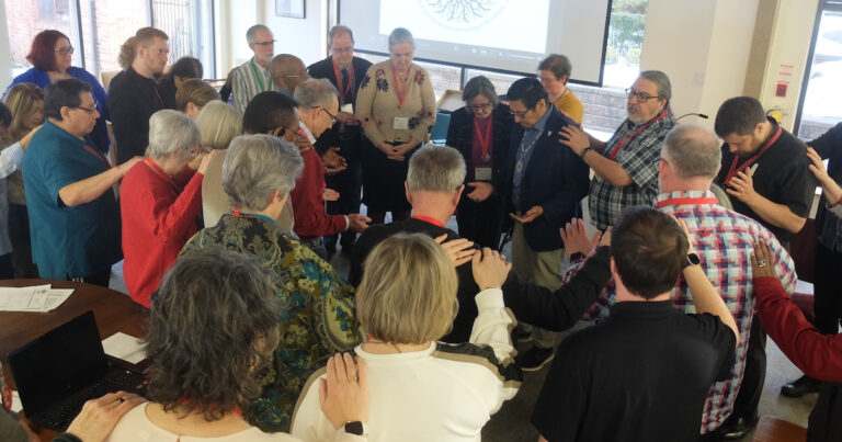 A diverse group of people gathered in a circle, with some individuals bowing their heads and others placing hands on shoulders, engaged in a moment of reflection or prayer during a community event.