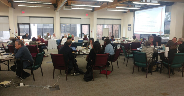 A group of professionals engaged in discussion and collaboration at a conference room, with tables arranged for teamwork and presentations.
