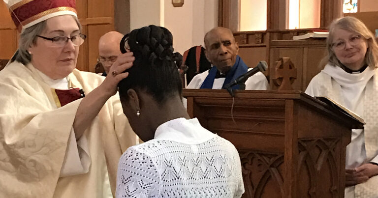 A young girl in a white dress is receiving a blessing during a religious ceremony, while two adults, one in a white robe and another in a blue scarf, observe attentively in a church setting.