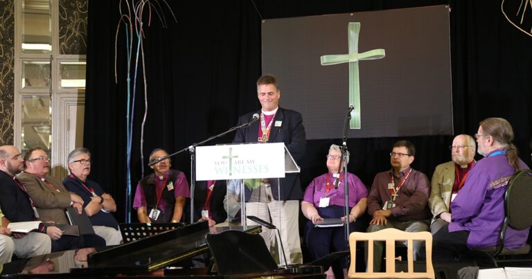 A speaker at a podium addresses an audience during a religious event, with a cross displayed in the background.