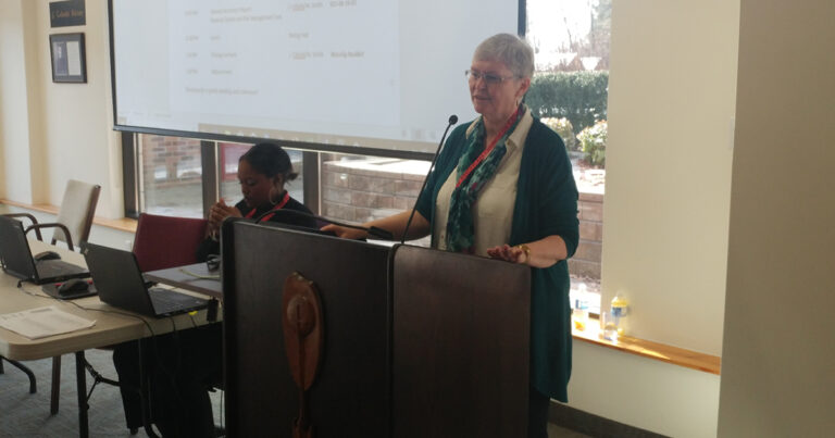A woman with short gray hair speaks at a podium during a conference, wearing a teal scarf and a black cardigan, while another person sits nearby.