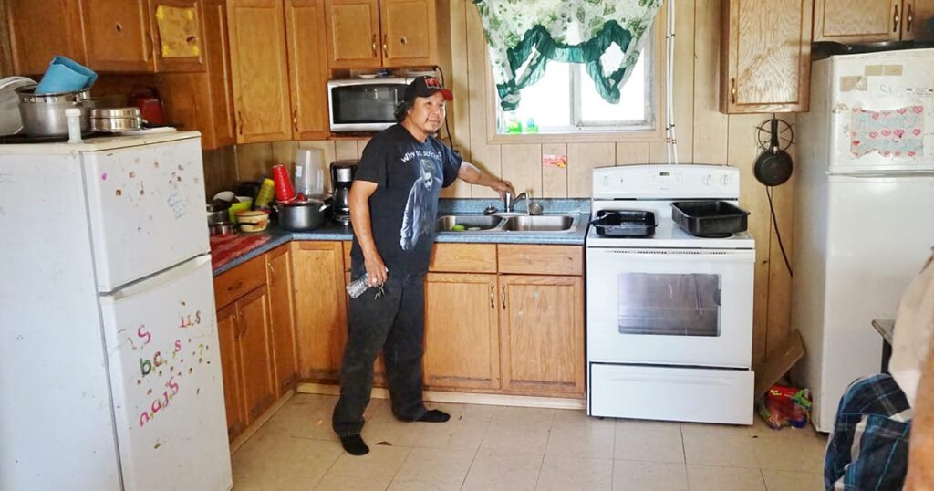 A man in a baseball cap stands in a kitchen beside the sink, demonstrating running water.