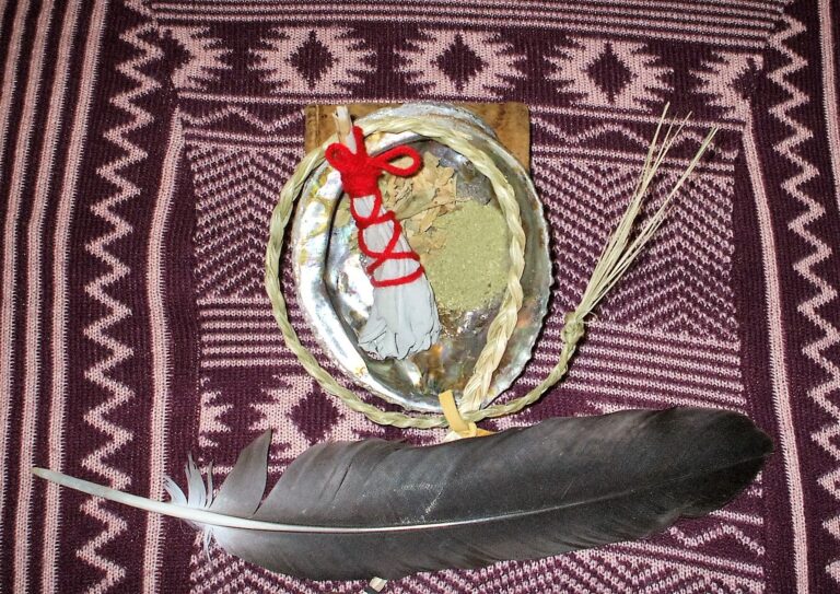 A handmade ceremonial pouch featuring a round shell adorned with red and white decorative elements, displayed on a patterned fabric background. A large feather is placed beside the pouch, emphasizing its cultural significance.