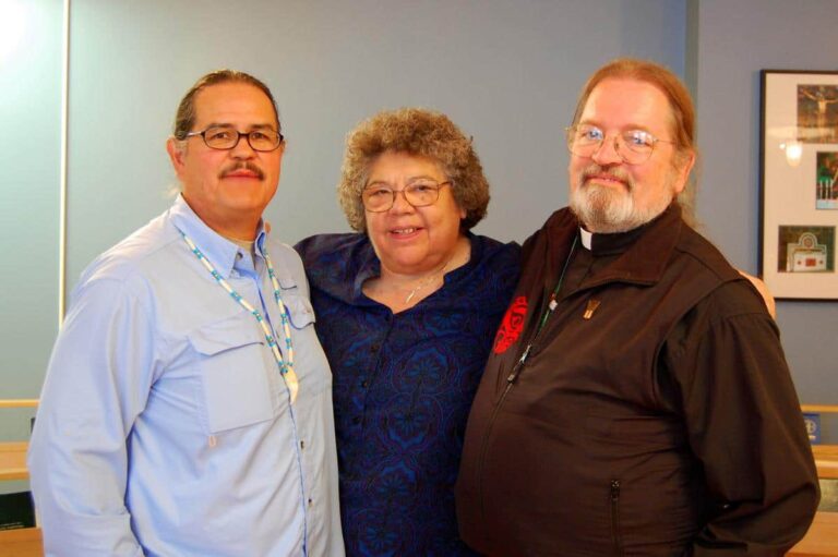 Three individuals standing together, smiling. The person on the left is wearing a light blue shirt with a necklace, the woman in the center is in a blue top, and the man on the right is wearing a dark jacket. The background is a soft blue color, creating a warm and inviting atmosphere.