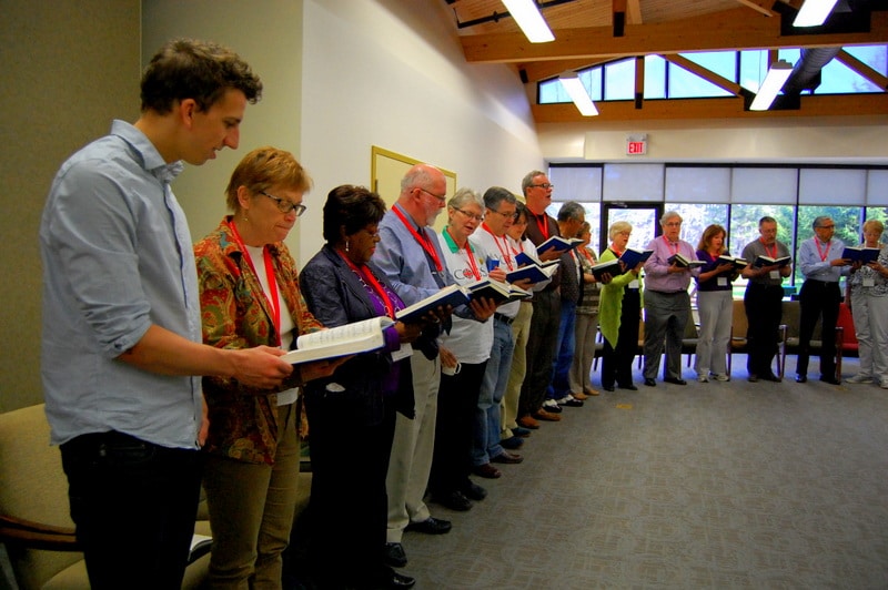 A diverse group of people standing in a line, holding music sheets and singing together in a brightly lit room with wooden beams.