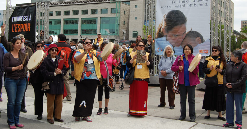 Participants in traditional attire perform a cultural celebration outdoors, holding drums and instruments, with a large banner visible in the background.