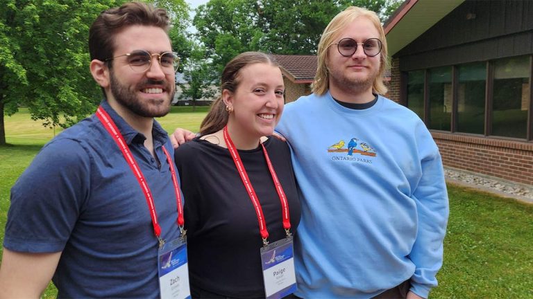 Three young white people stand outdoors with grass, trees and low buildings in the background. One is a young man with short brown hair, a beard and glasses. The second is a young woman with a brown pony tail. And the third his a taller young man with shoulder length blonde hair, a beard and glasses.