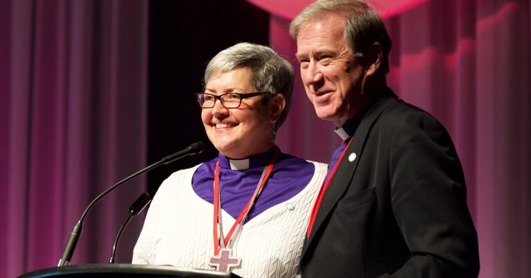 Two individuals stand together on stage, smiling at the audience during a formal event. One is wearing glasses and a white sweater with a purple collar, while the other is dressed in a black suit. Both are engaged in a moment of camaraderie, likely during a presentation or ceremony.