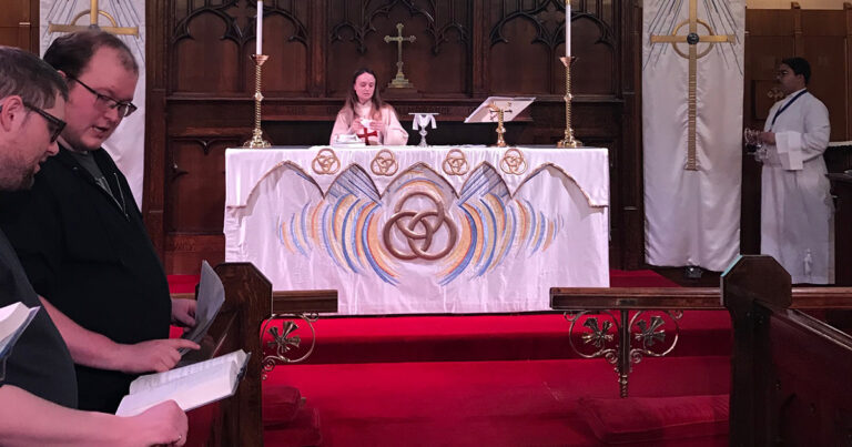 A woman standing at an altar in a church, with a decorated white altar cloth featuring a trinity symbol, surrounded by candles and a red carpet.