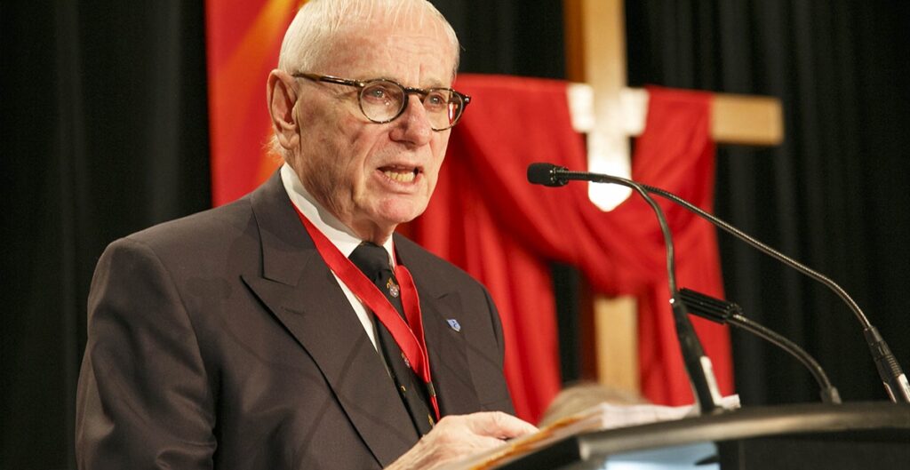 Elderly man wearing glasses and a suit, speaking at a podium with a microphone, set against a backdrop of red drapery.
