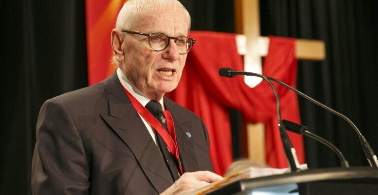 Elderly man wearing glasses and a suit, speaking at a podium with a microphone, set against a backdrop of red drapery.