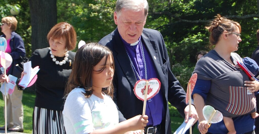 A man in a clergy outfit interacts with a young girl holding a heart-shaped craft, both smiling in a sunny outdoor setting during a community event.