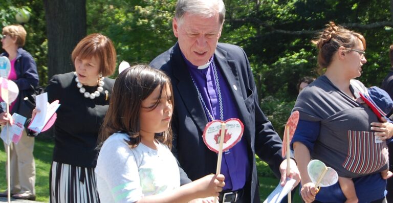 A man in a clergy outfit interacts with a young girl holding a heart-shaped craft, both smiling in a sunny outdoor setting during a community event.