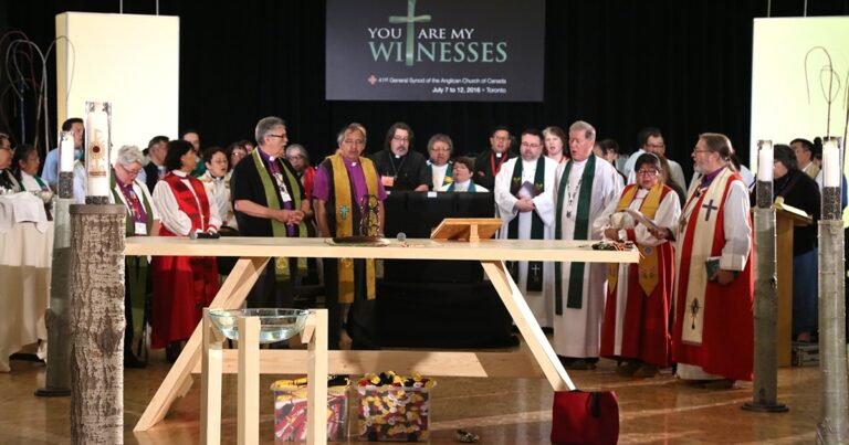A diverse group of clergy members gathered around a wooden altar during a religious ceremony, with a backdrop displaying the text "You Are My Witnesses." The setting conveys a sense of community and spiritual significance.