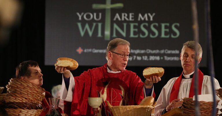 A priest in red vestments holds two loaves of bread during a religious ceremony, with a backdrop displaying the text "You Are My Witnesses." The scene emphasizes the significance of communion in a church setting.