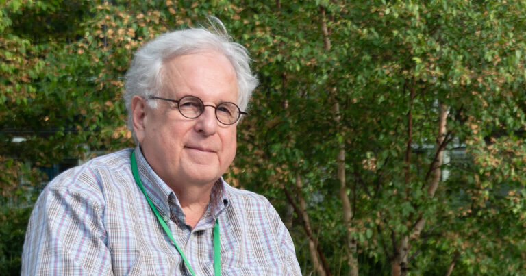 An older man with glasses and gray hair is smiling while wearing a plaid shirt and a green lanyard, surrounded by greenery.