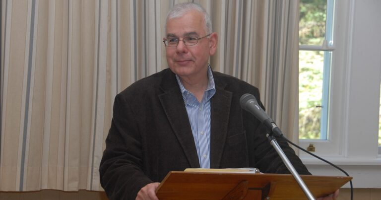 A middle-aged man with glasses stands at a wooden podium, wearing a dark blazer and light blue shirt, speaking into a microphone. The background features soft, neutral-colored drapery.