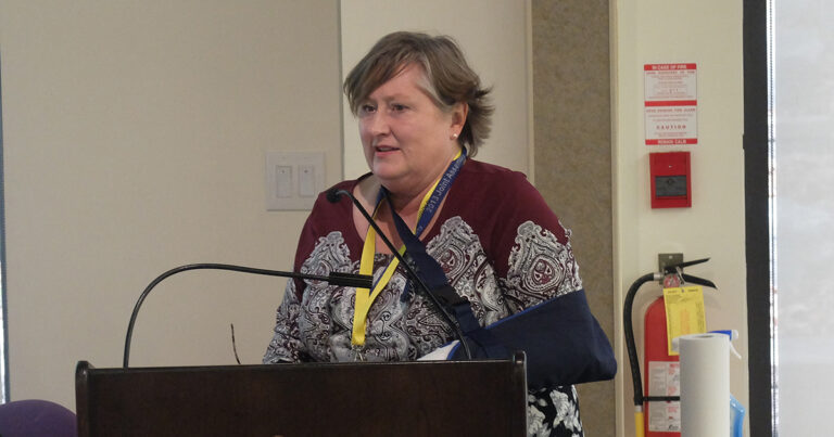 A woman with short brown hair, wearing a patterned sweater, speaks at a podium with a microphone, showcasing a presentation at a conference.