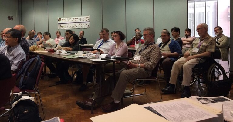 A diverse group of participants attentively listening during a workshop or meeting at a conference, seated around a table with notebooks and materials in front of them.