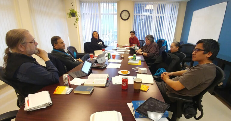 Members of the Anglican Council of Indigenous Peoples sit around a conference table in discussion, with laptops, notebooks, and coffee cups spread across the table.