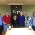 Group of six people standing together in a well-lit room with a table in front, featuring two candles. They are dressed in a mix of casual and formal attire, with a smiling woman in the center and a man wearing a purple shirt. The background shows a decorative door and green walls.