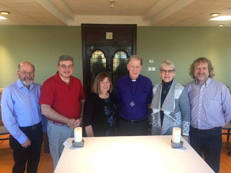 Group of six people standing together in a well-lit room with a table in front, featuring two candles. They are dressed in a mix of casual and formal attire, with a smiling woman in the center and a man wearing a purple shirt. The background shows a decorative door and green walls.