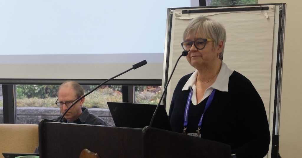 Woman speaking at a podium during a meeting while another person sits at a laptop in the background near a presentation screen