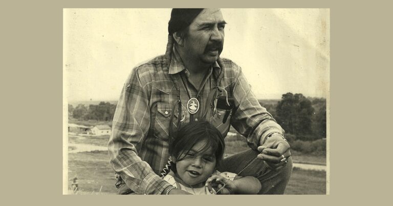 A black and white photograph of a man wearing a plaid shirt and a necklace, sitting outdoors with a young girl who is smiling and leaning against him. The background features a grassy landscape.