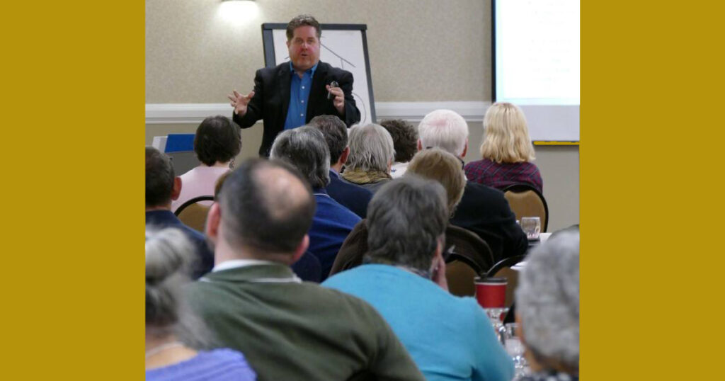A speaker presenting to an audience in a conference setting, with attendees engaged and seated at tables.