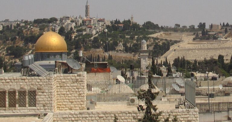 Panoramic view of the Old City of Jerusalem, featuring the iconic golden dome of the Dome of the Rock, surrounded by ancient stone walls and lush greenery in the background.