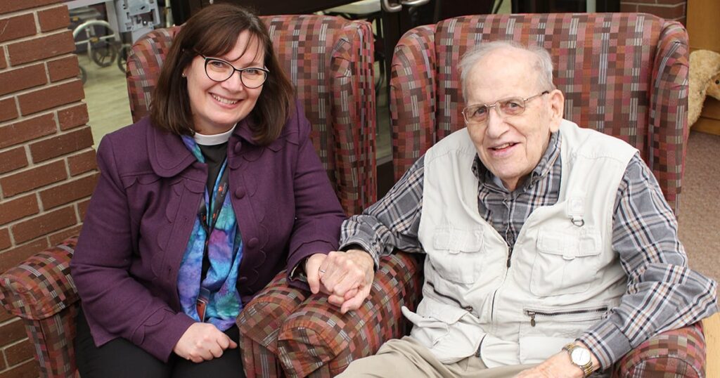 A smiling woman and an elderly man sit together on a couch, holding hands, in a warm and inviting setting. The woman wears glasses and a colorful top, while the man is dressed in a light vest over a checkered shirt.