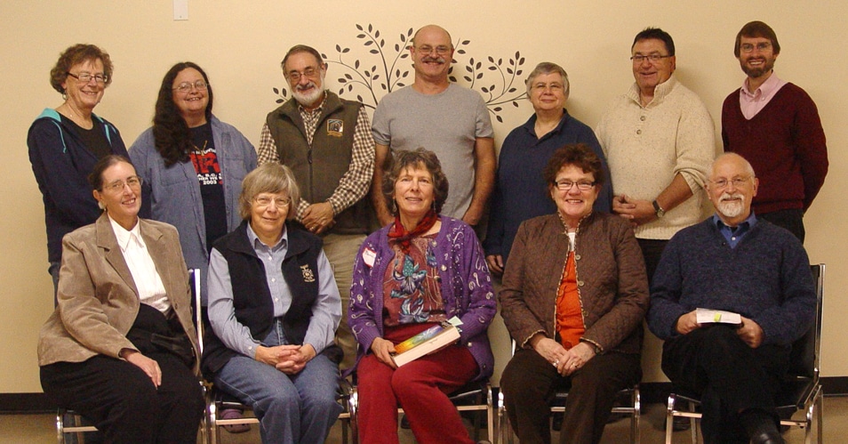 Group photo of ten individuals smiling together, seated and standing, in a community setting with a decorative wall in the background.