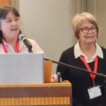 Two women speaking at a conference, one at a podium with a microphone and a laptop, while the other stands beside her, both wearing red lanyards.