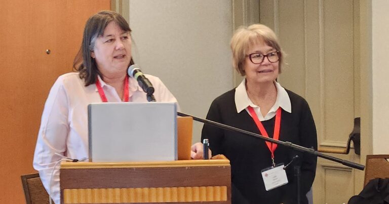 Two women speaking at a conference, one at a podium with a microphone and a laptop, while the other stands beside her, both wearing red lanyards.